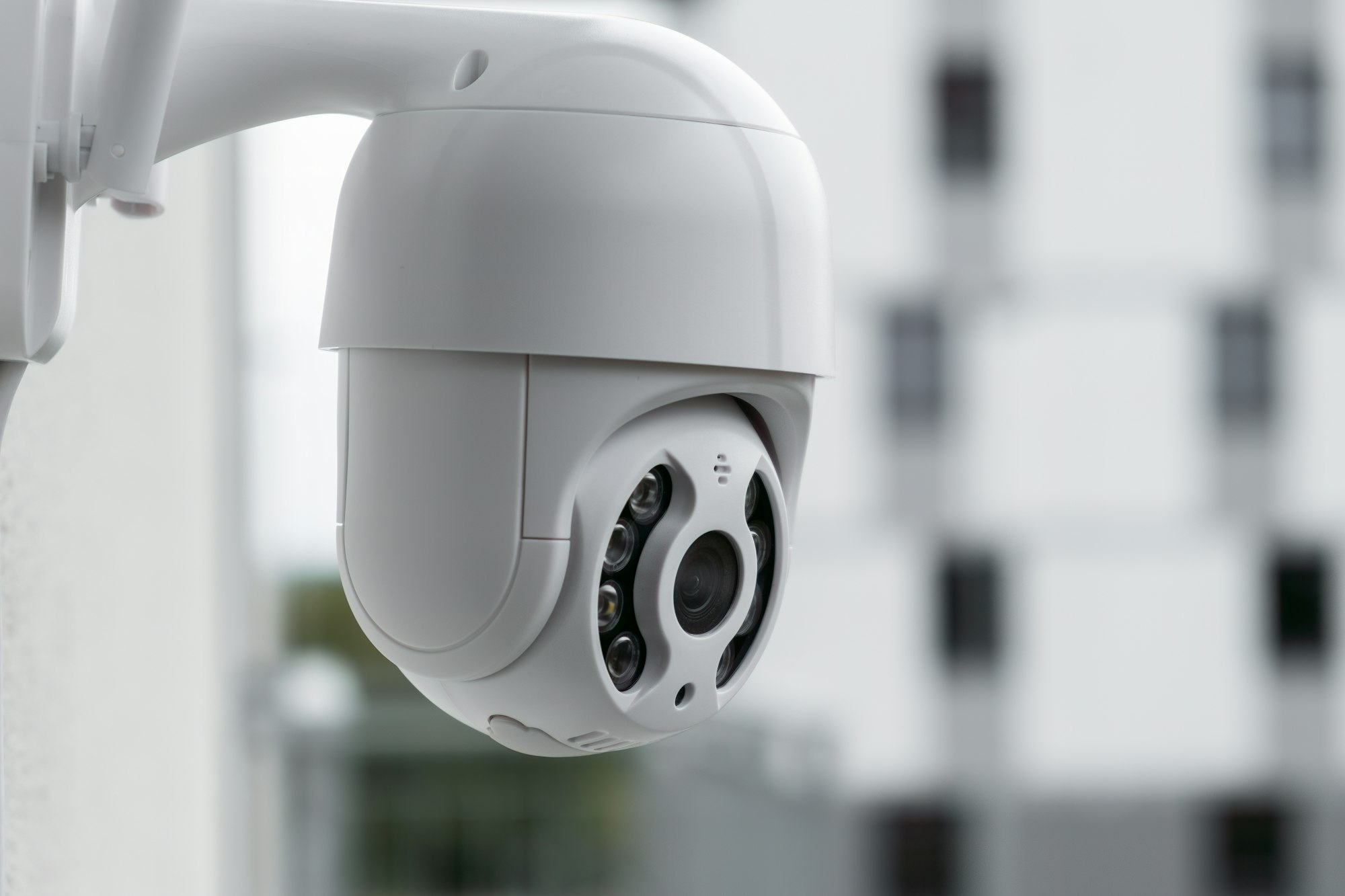 Modern CCTV camera on a wall with a blurred multi-storey building on the background.